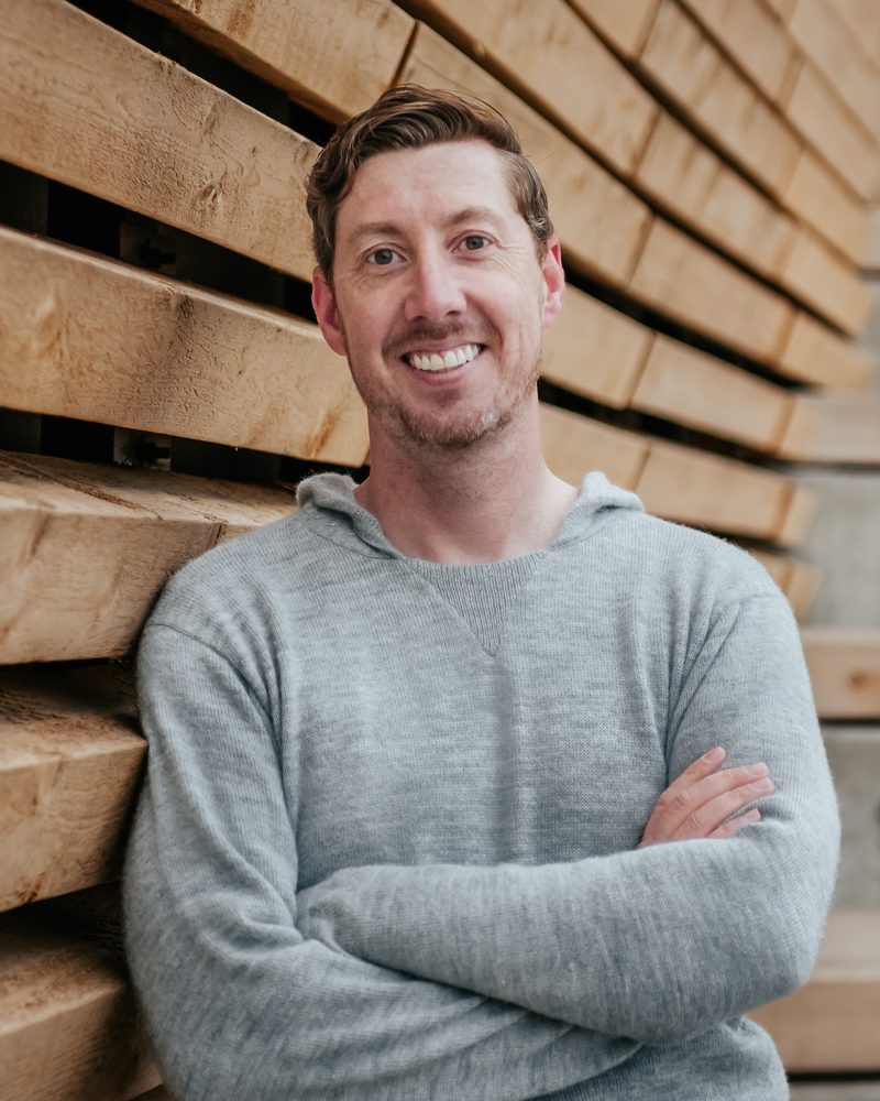 Kyle Tabor, smiling with arms crossed, leaning against a wall of stacked timber